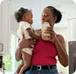 Woman holding a child and drinking a smoothie in a bright kitchen.